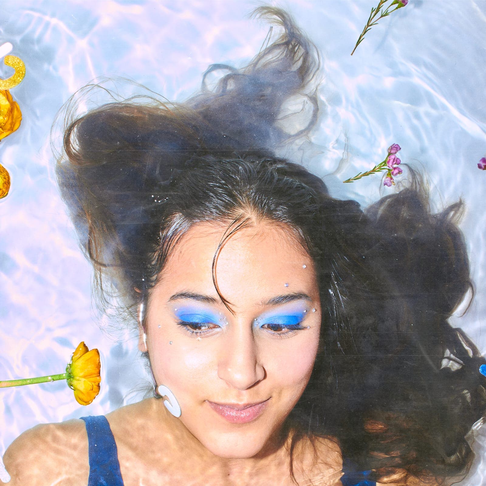 Qiwi Chee allongée dans une eau claire, les cheveux déployés autour du visage, fard à paupières bleu vif, des fleurs jaunes et roses flottant à la surface.