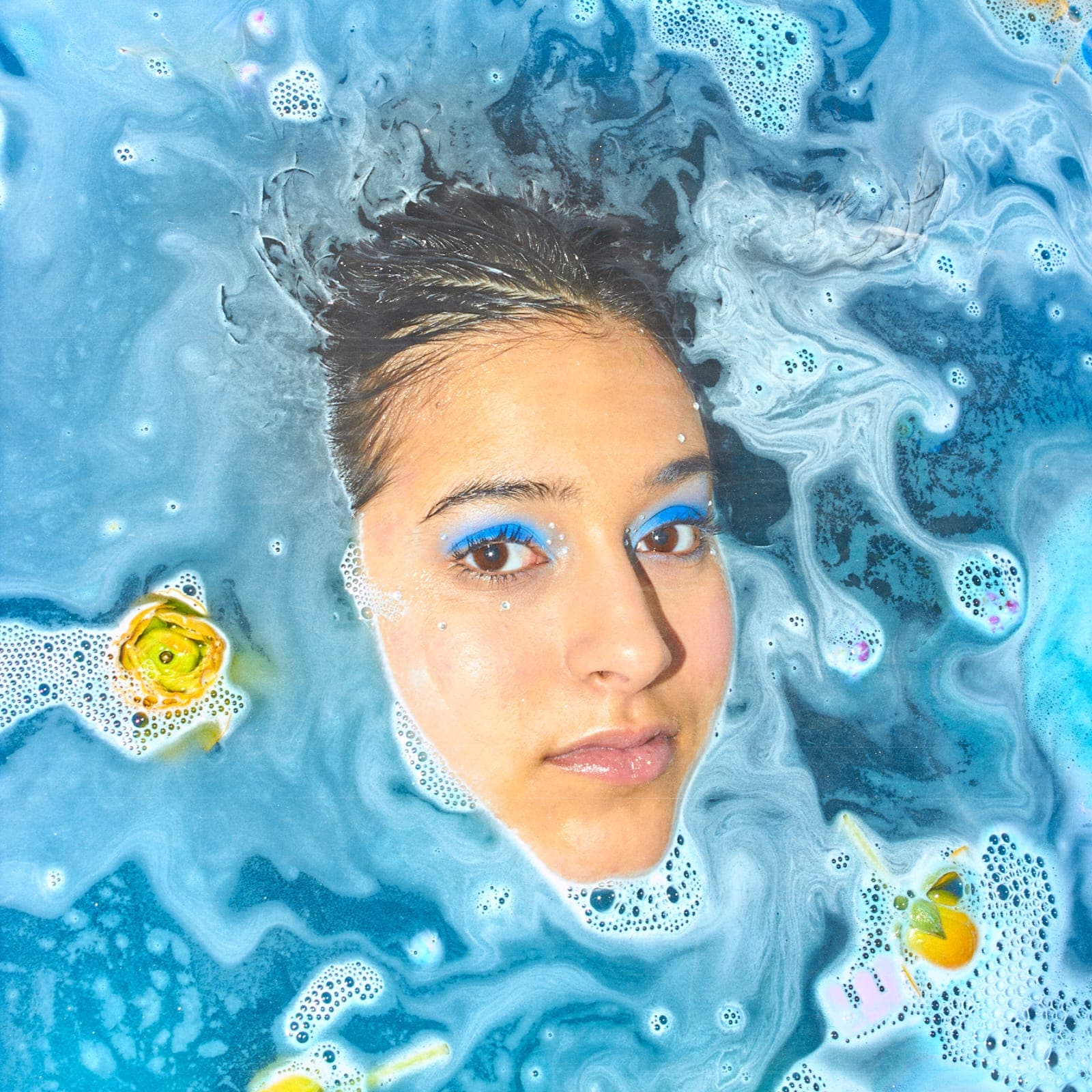 Le visage de Qiwi Chee émergeant d’une eau bleue couverte de mousse, le regard tourné vers l’objectif, deux fleurs jaunes flottant de part et d’autre.