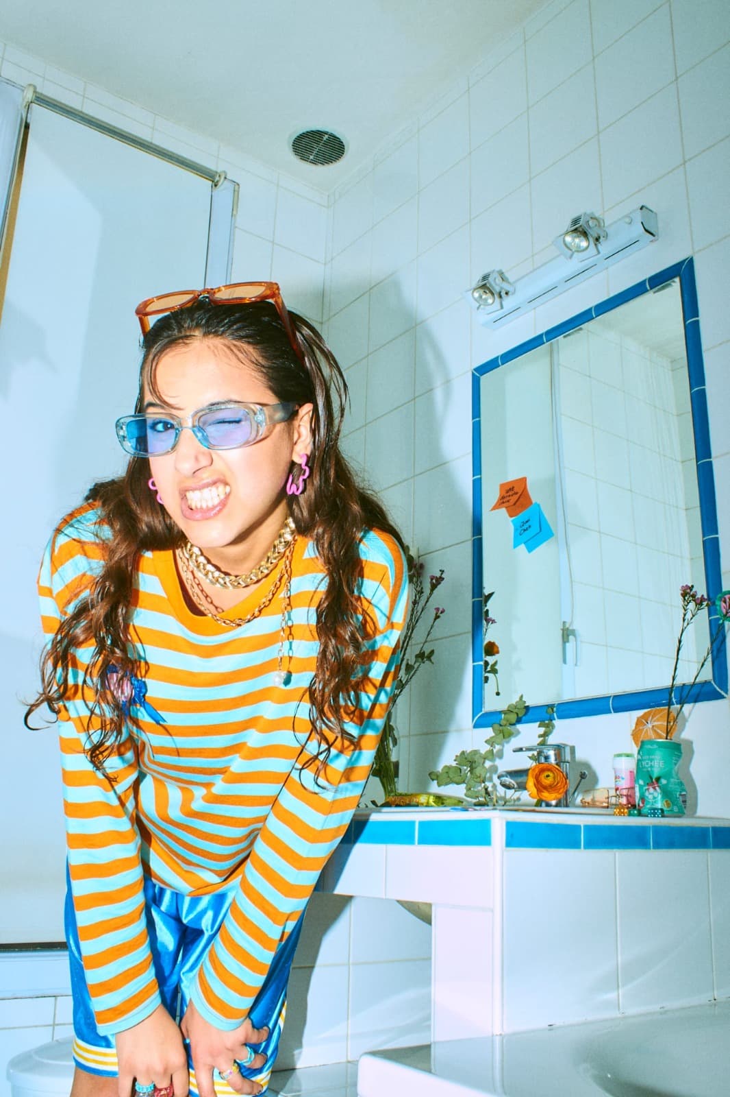 Qiwi Chee penchée en avant, lunettes bleues sur le nez et lunettes orange relevées sur la tête, tirant la langue à l’objectif.